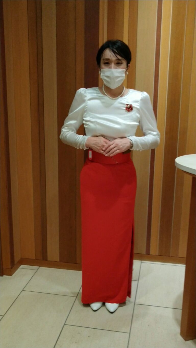 ホテルの木目壁前に立つ白ブラウスと赤いロングスカートのバンケットコンパニオン女装